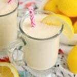 Front view of a chilled glass of frosted lemonade with a lemon slice on the rim and a pink straw, surrounded by fresh lemon wedges.