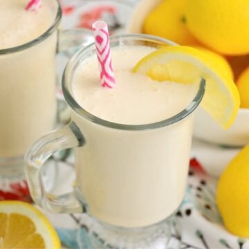 Front view of a chilled glass of frosted lemonade with a lemon slice on the rim and a pink straw, surrounded by fresh lemon wedges.