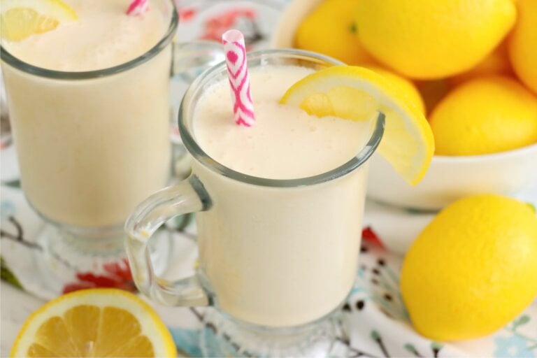 Front view of a chilled glass of frosted lemonade with a lemon slice on the rim and a pink straw, surrounded by fresh lemon wedges.