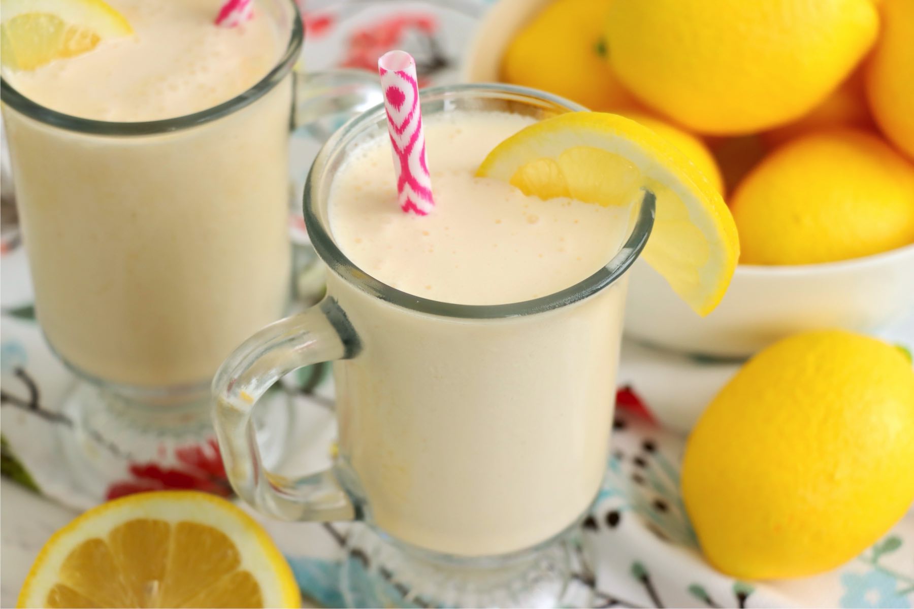 Front view of a chilled glass of frosted lemonade with a lemon slice on the rim and a pink straw, surrounded by fresh lemon wedges.