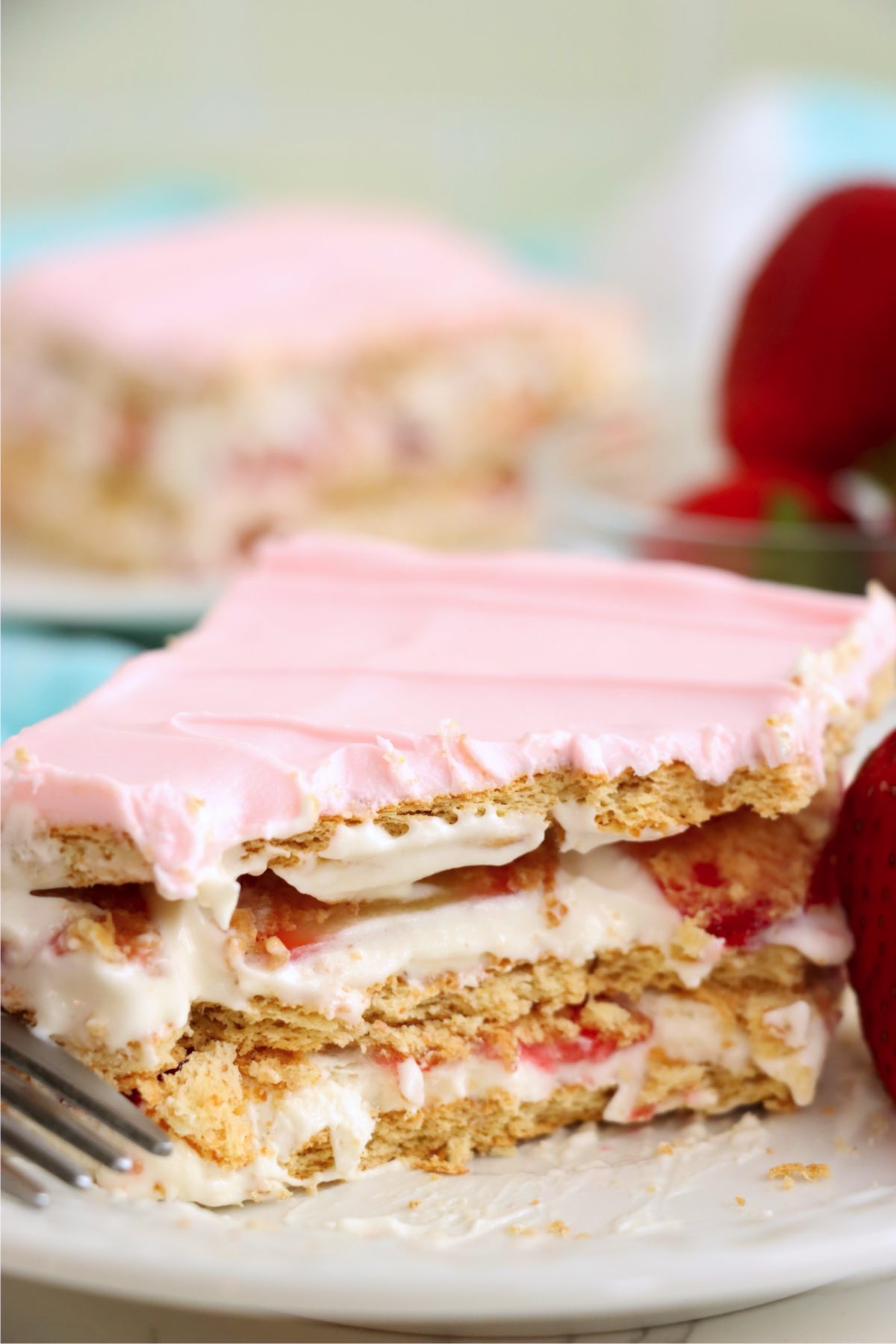 A close-up of a dessert slice showing layers of graham crackers, creamy filling, sliced strawberries, and topped with pale pink frosting, with a fork resting on the plate.