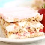 A slice of dessert on a white plate, clearly revealing its layers of graham crackers, creamy pudding, sliced strawberries, and pink frosting, garnished with a fresh strawberry.