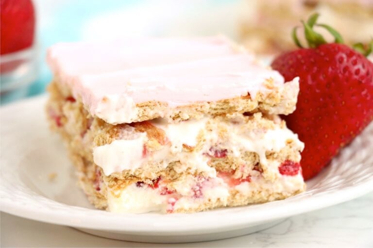 A slice of dessert on a white plate, clearly revealing its layers of graham crackers, creamy pudding, sliced strawberries, and pink frosting, garnished with a fresh strawberry.