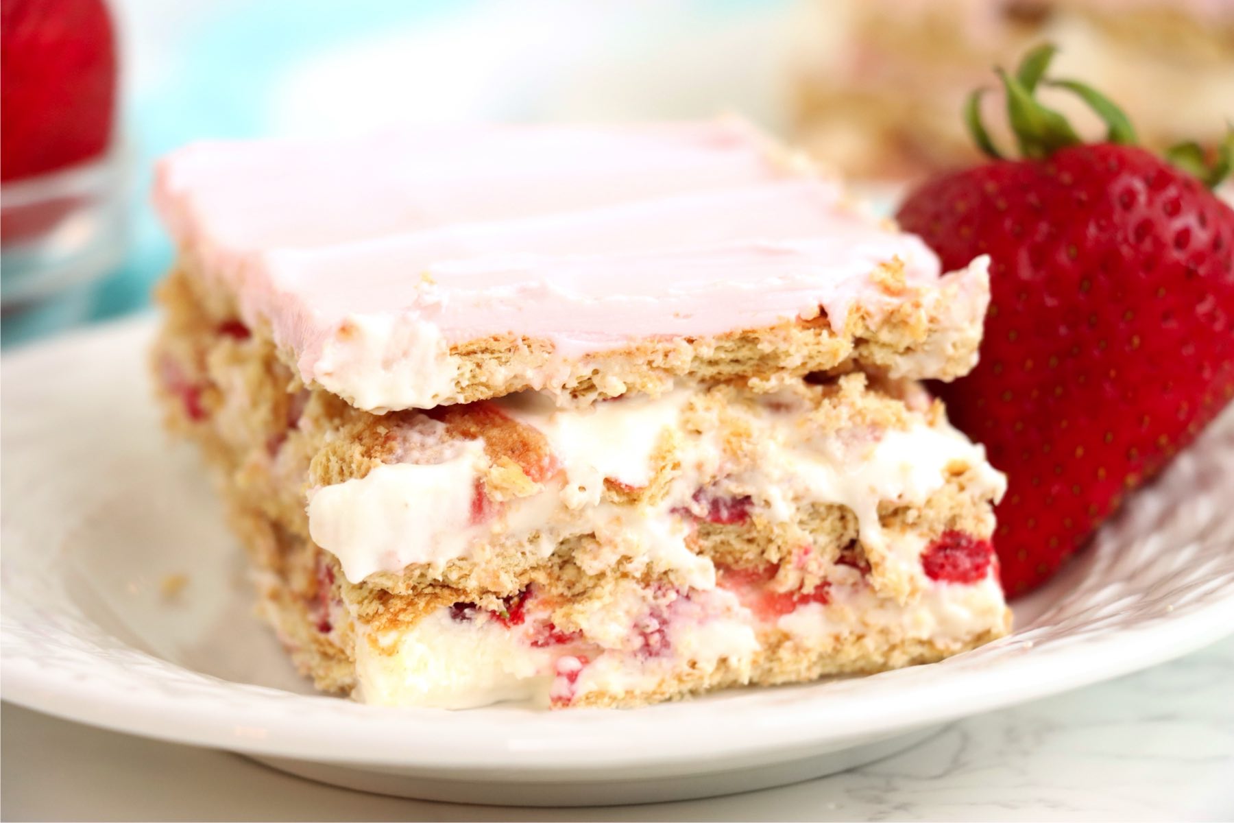 A slice of dessert on a white plate, clearly revealing its layers of graham crackers, creamy pudding, sliced strawberries, and pink frosting, garnished with a fresh strawberry.