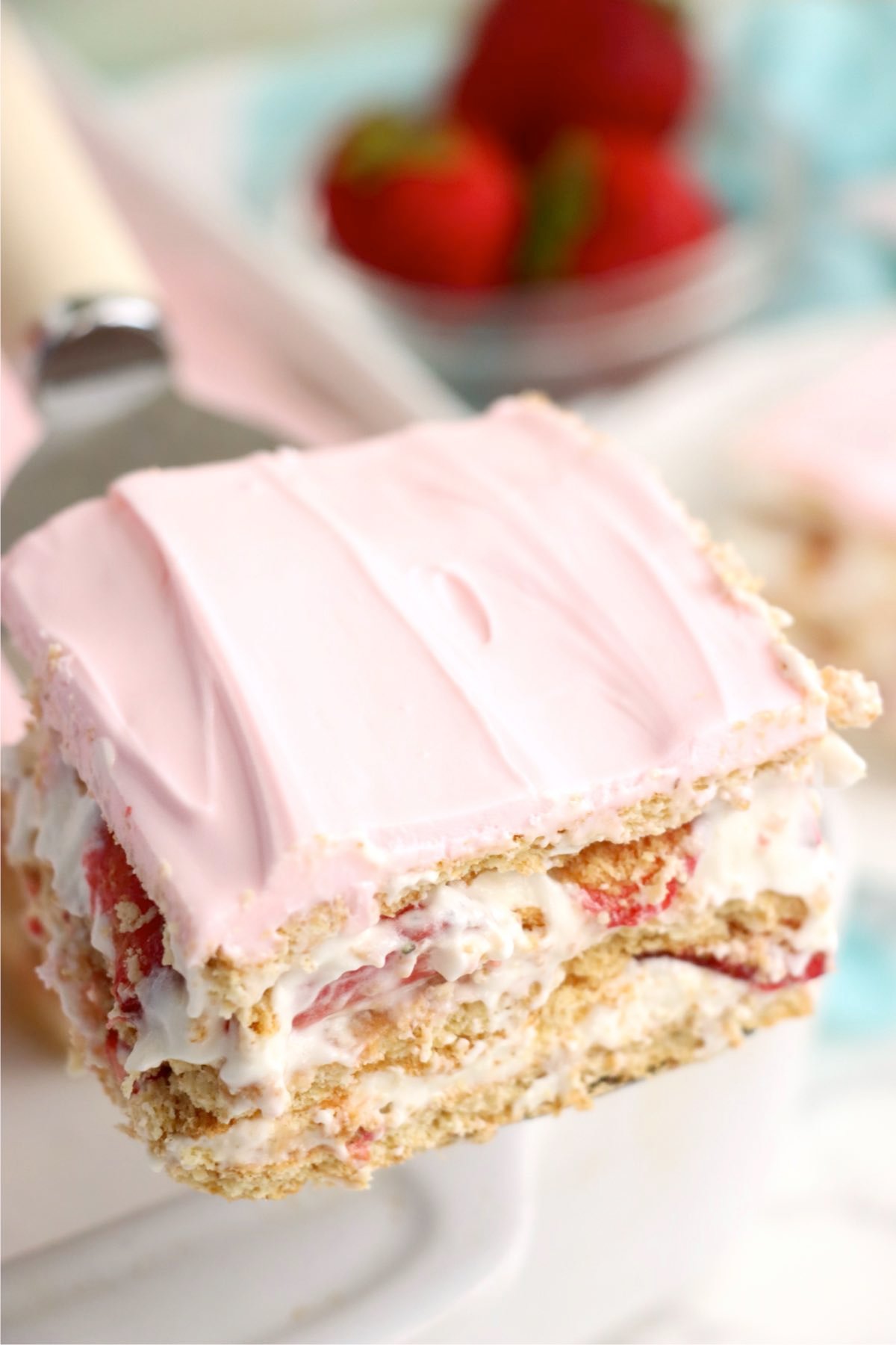 A serving spatula lifting a slice of dessert clearly showing layers of graham crackers, creamy filling, fresh strawberries, and topped with smooth, pale pink frosting.