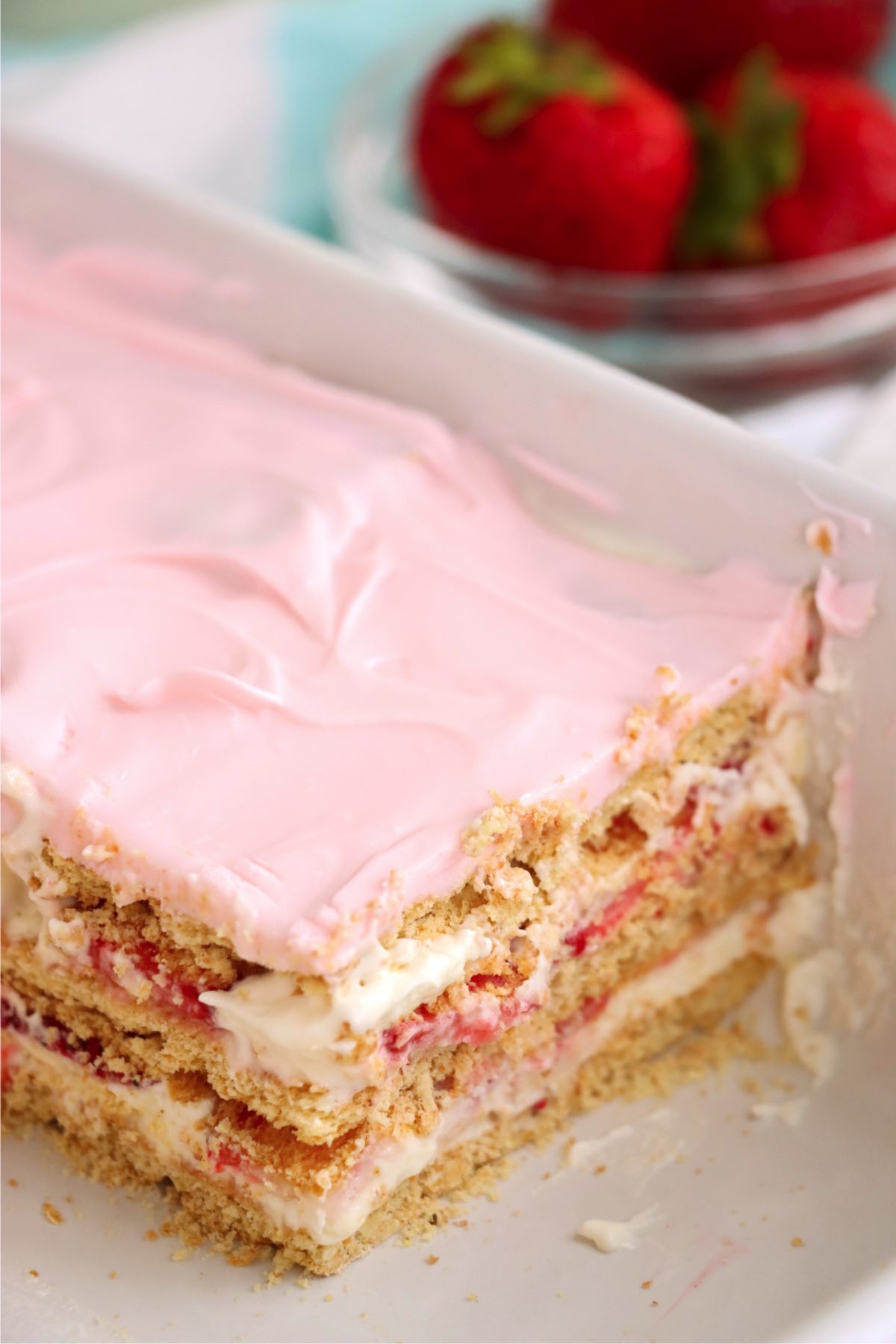 A view of the dessert in a baking dish after serving, revealing neatly layered graham crackers, creamy filling, strawberries, and pink frosting, alongside fresh strawberries in the background.