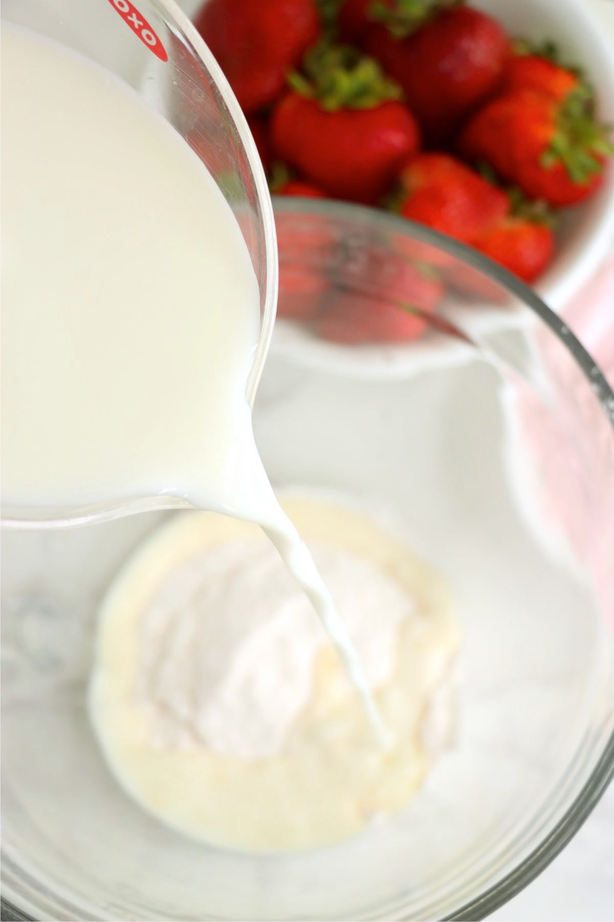Milk being poured from a measuring cup into a glass mixing bowl containing a powdered ingredient, with fresh strawberries nearby.