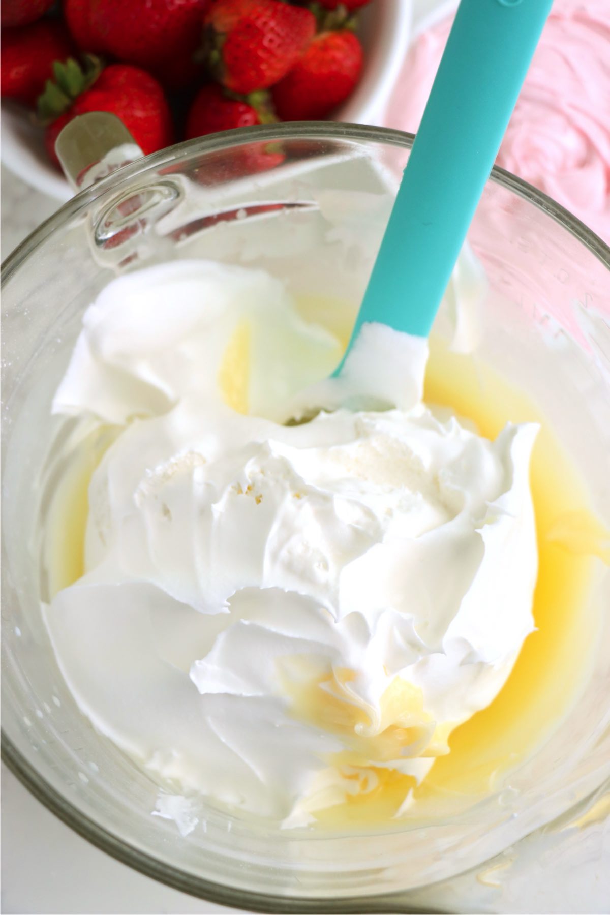 Whipped topping being folded gently into creamy pudding in a mixing bowl, with fresh strawberries in the background.