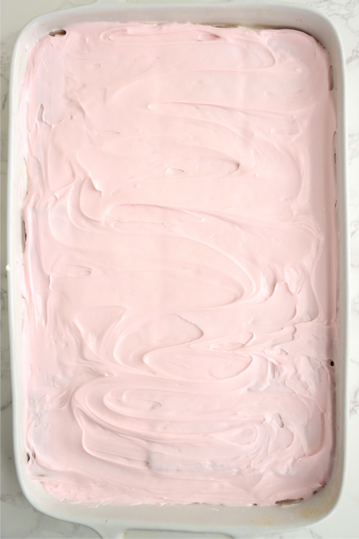 Soft pink frosting spread evenly across the top of the dessert, covering the final layer of graham crackers in a white rectangular baking dish.