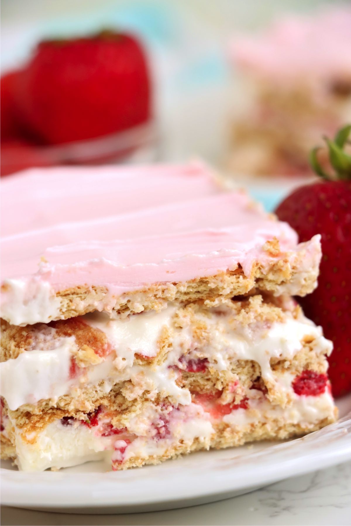 A close-up slice of the dessert showcasing layers of graham crackers, fresh strawberries, creamy pudding, and a soft pink frosting, served on a white plate with a whole strawberry beside it.