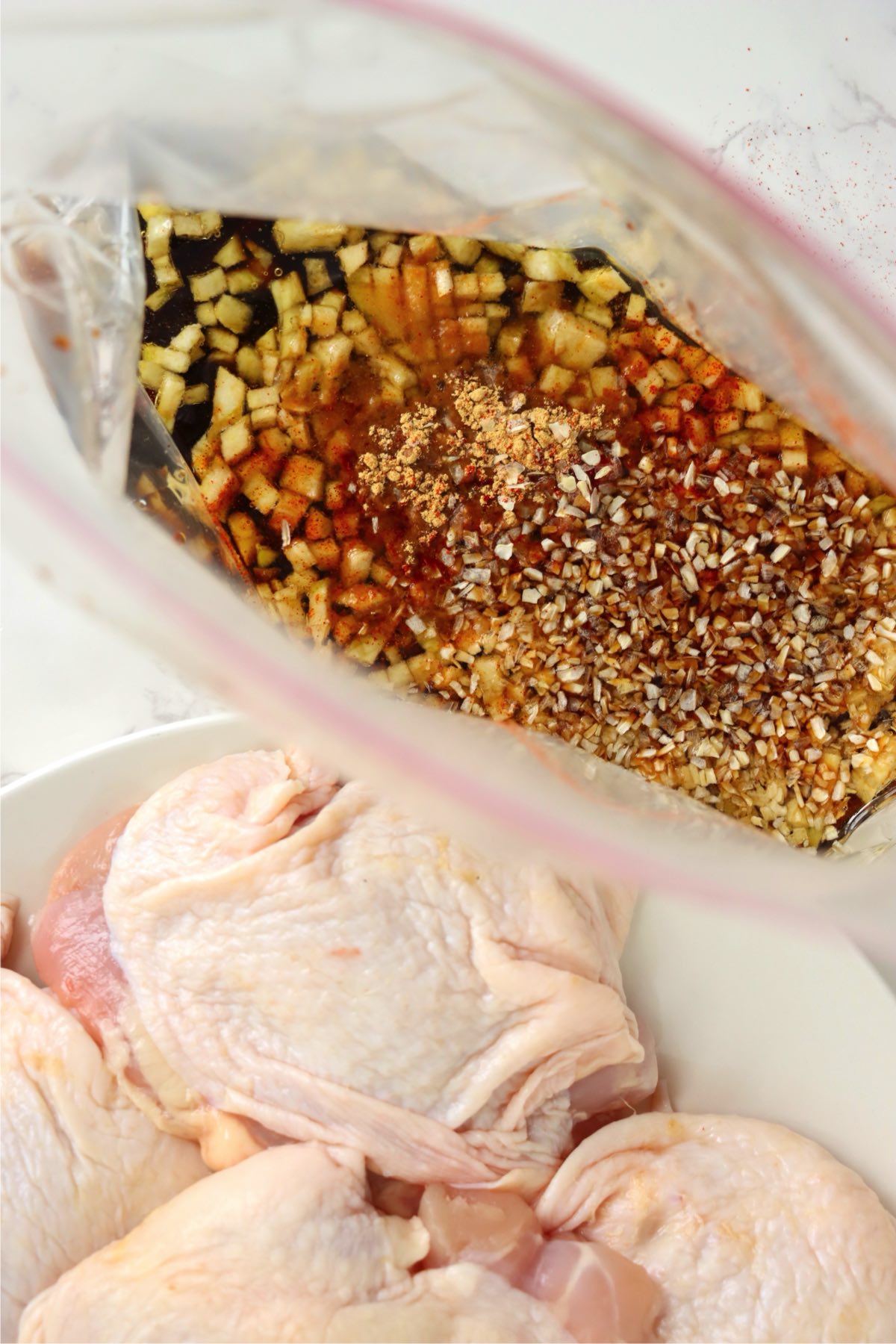 A close-up of marinade ingredients, including chopped garlic and onion, inside a ziplock bag next to raw chicken thighs in a bowl.