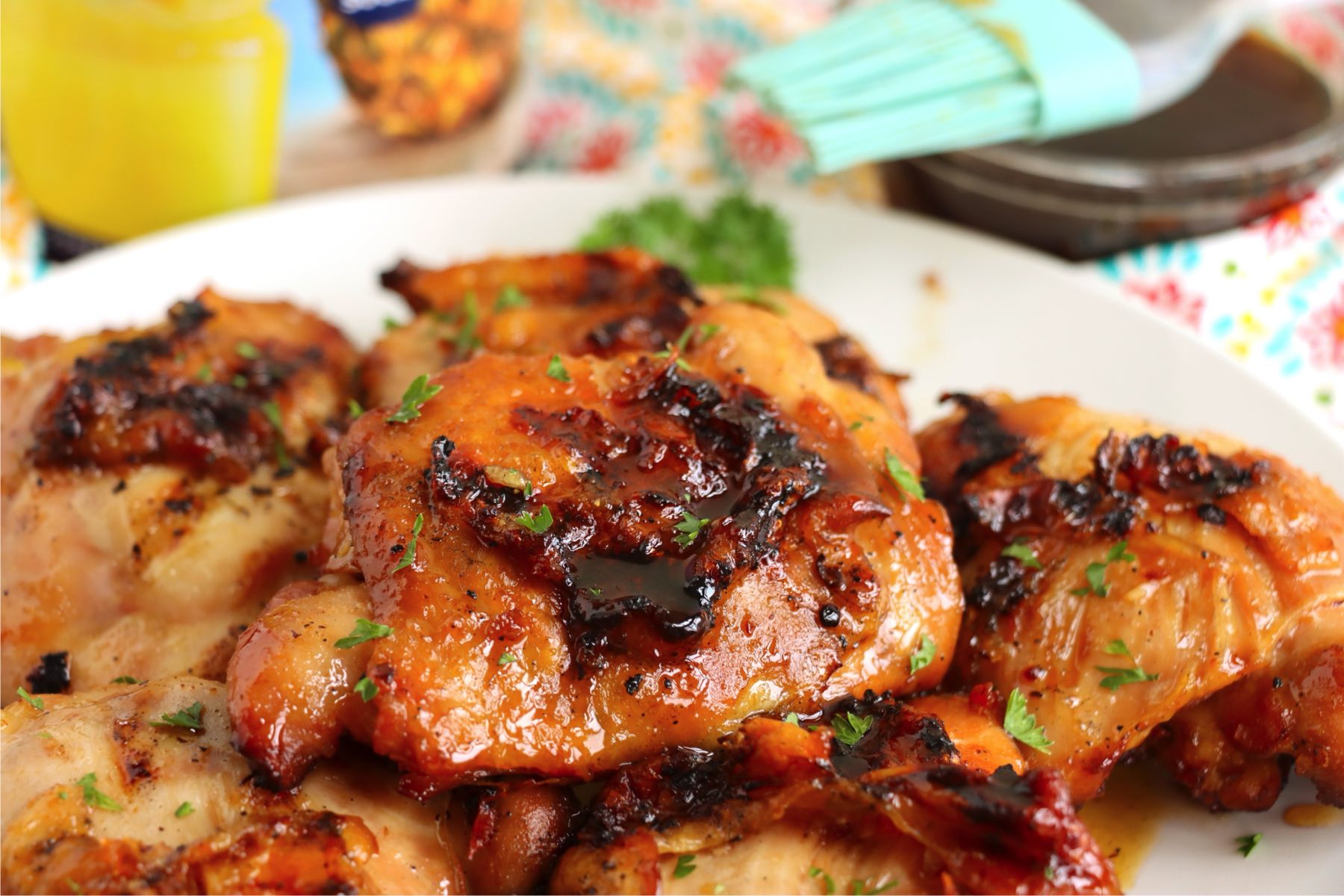 Grilled chicken thighs on a serving plate, with a marinade brush and pineapple juice container in the background.