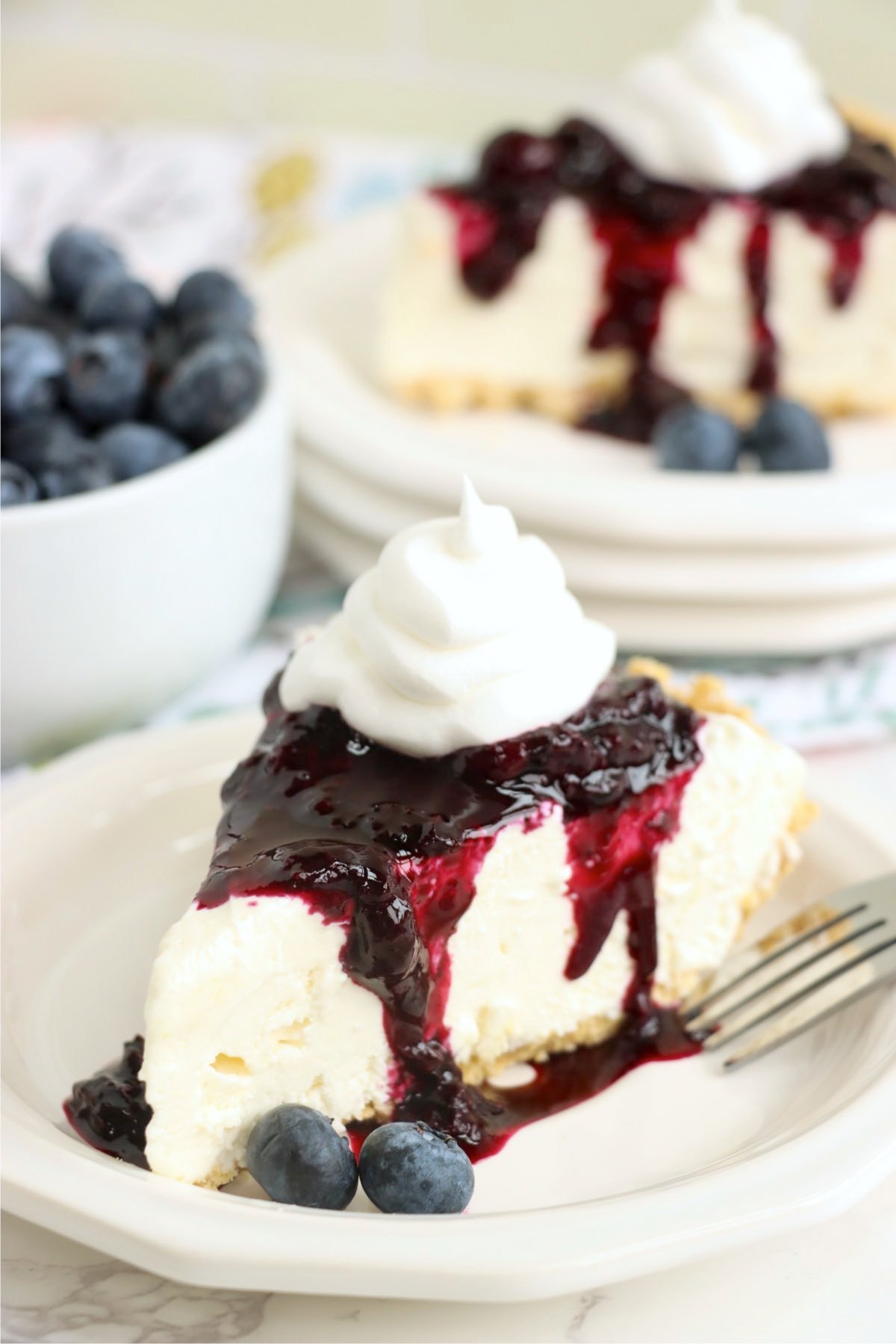 A slice of pie with blueberry sauce and whipped topping, with a few fresh blueberries on the plate.