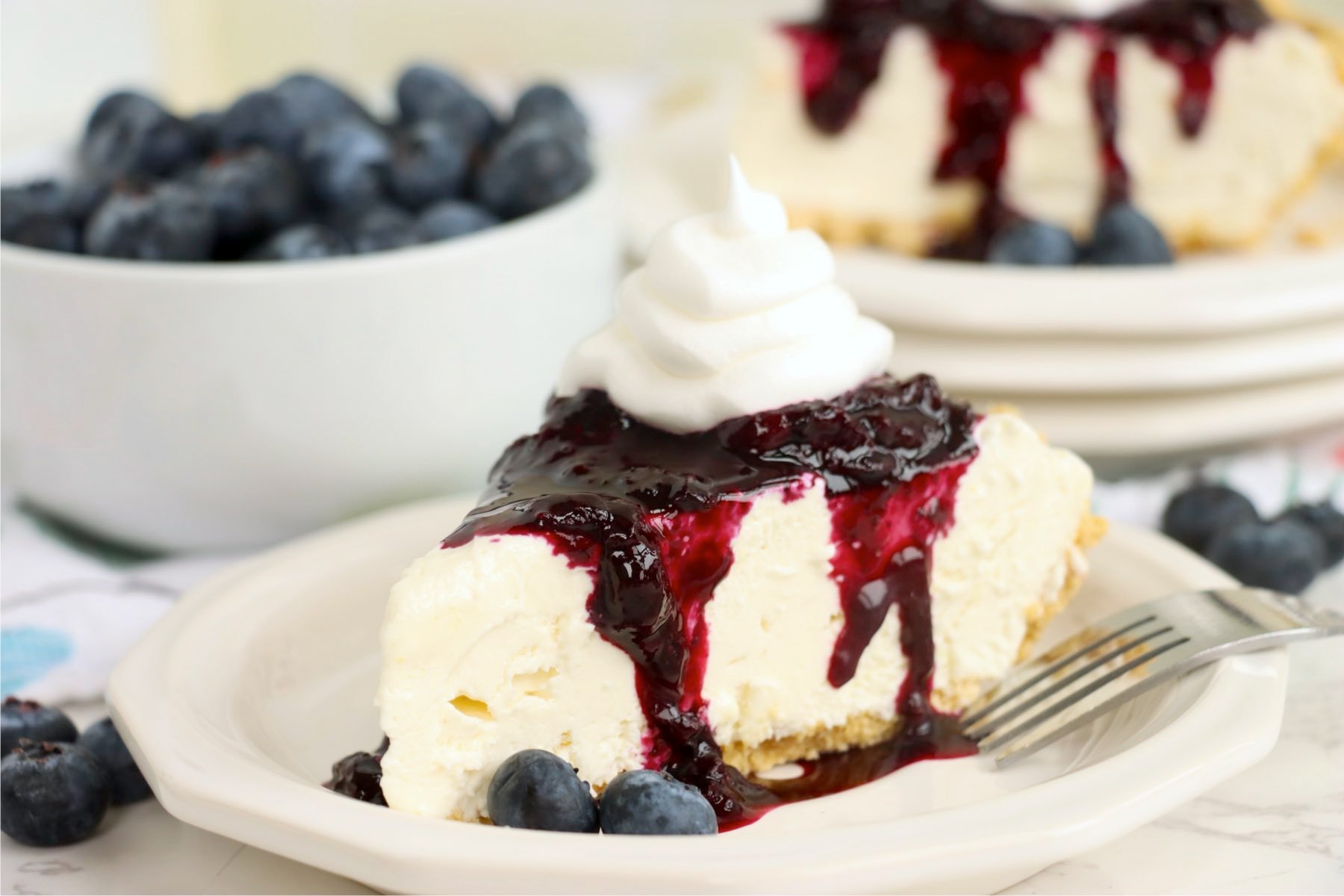 A slice of pie with blueberry sauce and whipped topping next to a bowl of fresh blueberries.