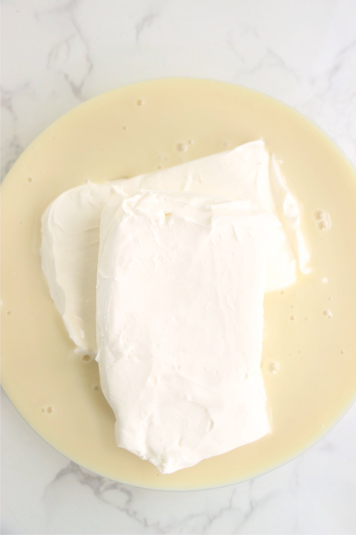 Two blocks of cream cheese sitting in a bowl of sweetened condensed milk.