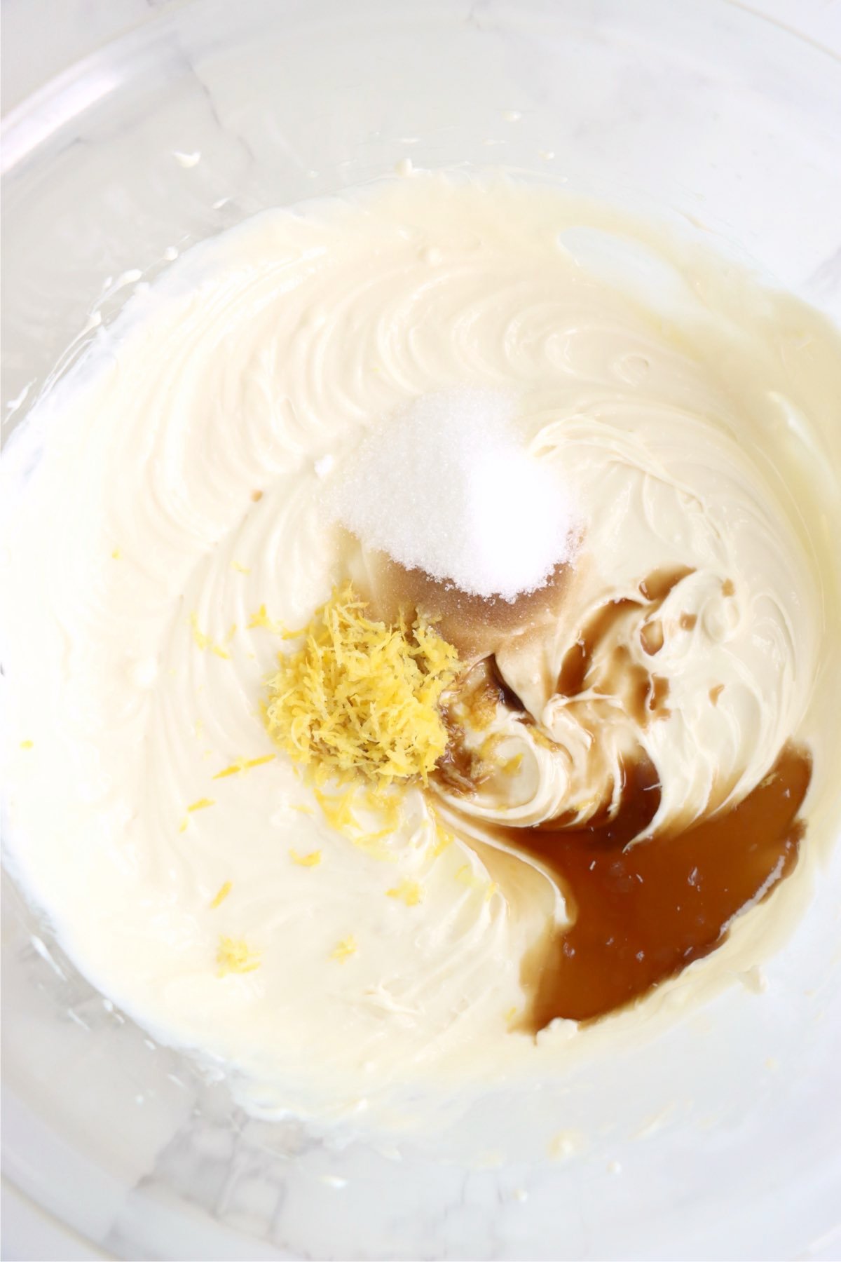 A mixing bowl with cream cheese mixture, lemon zest, sugar, and vanilla on top.