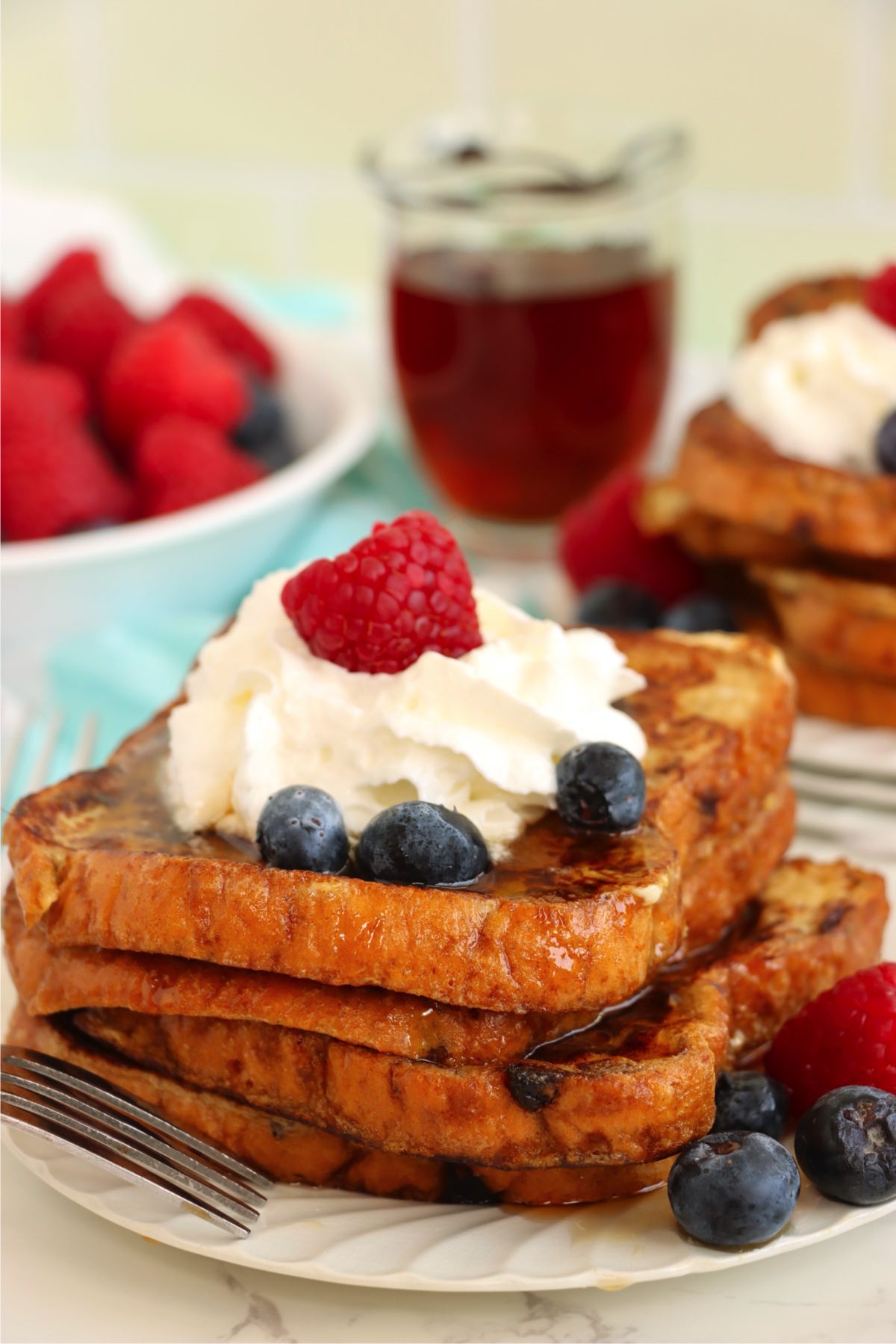 Plate of French toast stacked with whipped cream, blueberries, and a raspberry, with syrup and berries in the background.
