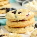 A stack of blueberry cookies on a plate surrounded by scattered blueberries and white chocolate chips.