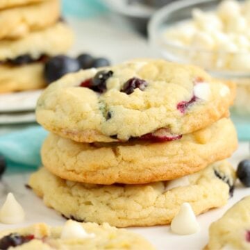 A stack of blueberry cookies on a plate surrounded by scattered blueberries and white chocolate chips.
