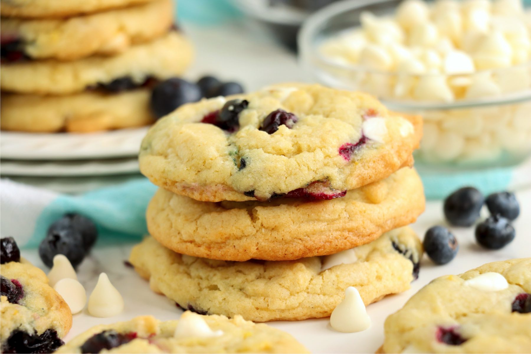 A stack of blueberry cookies on a plate surrounded by scattered blueberries and white chocolate chips.