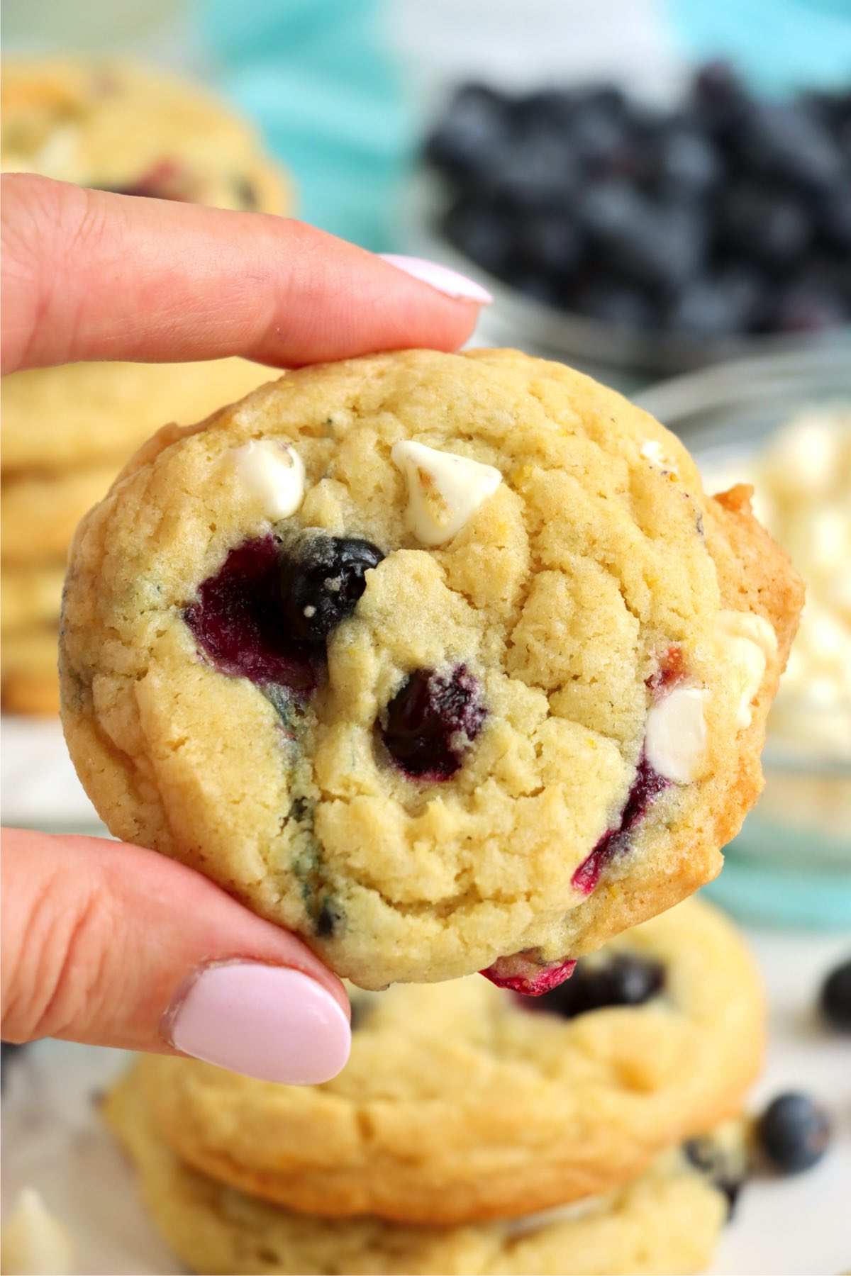 A hand holding a single blueberry cookie to show its golden texture and mix of blueberries and white chocolate chips.