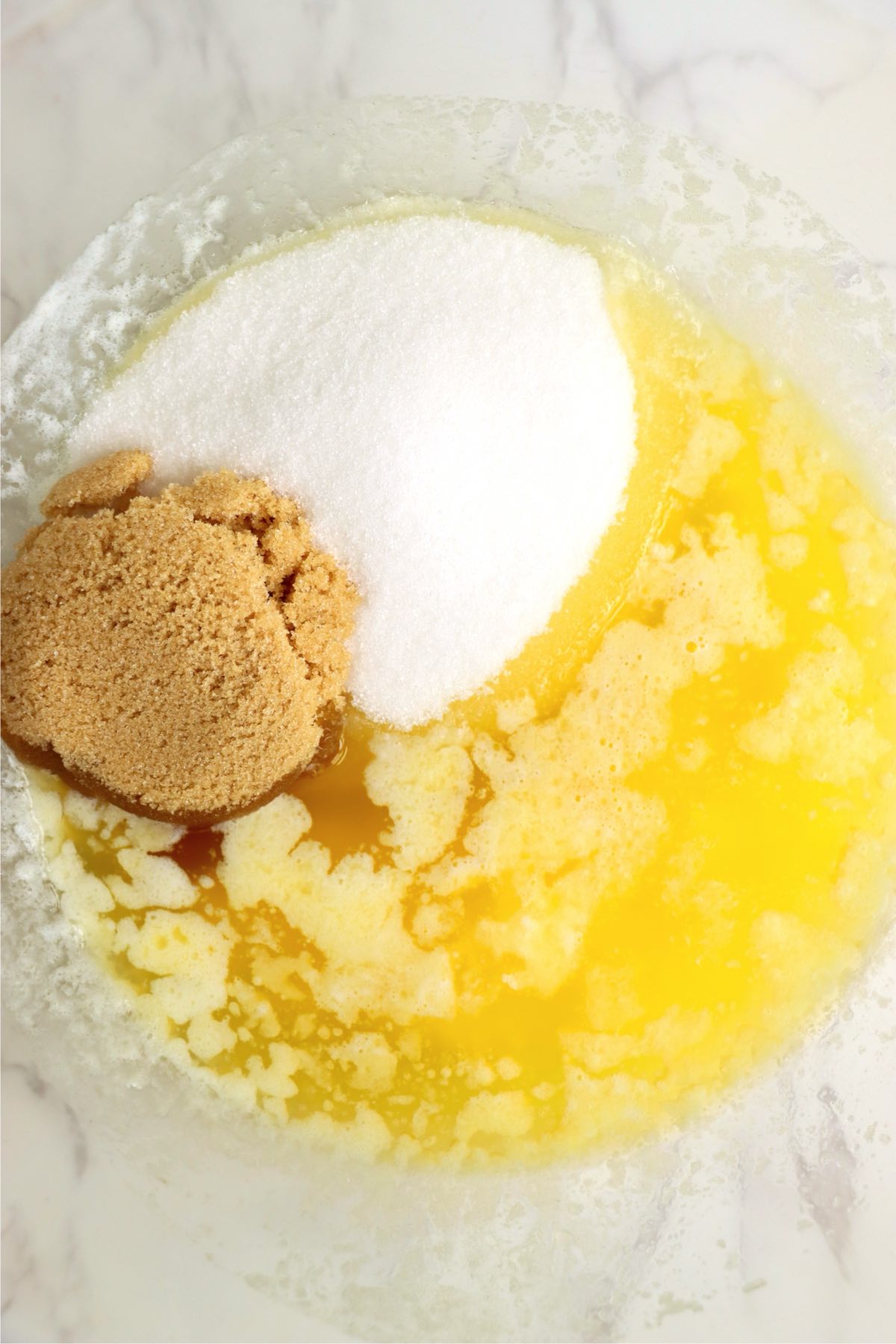Melted butter in a mixing bowl with white sugar and brown sugar on top.