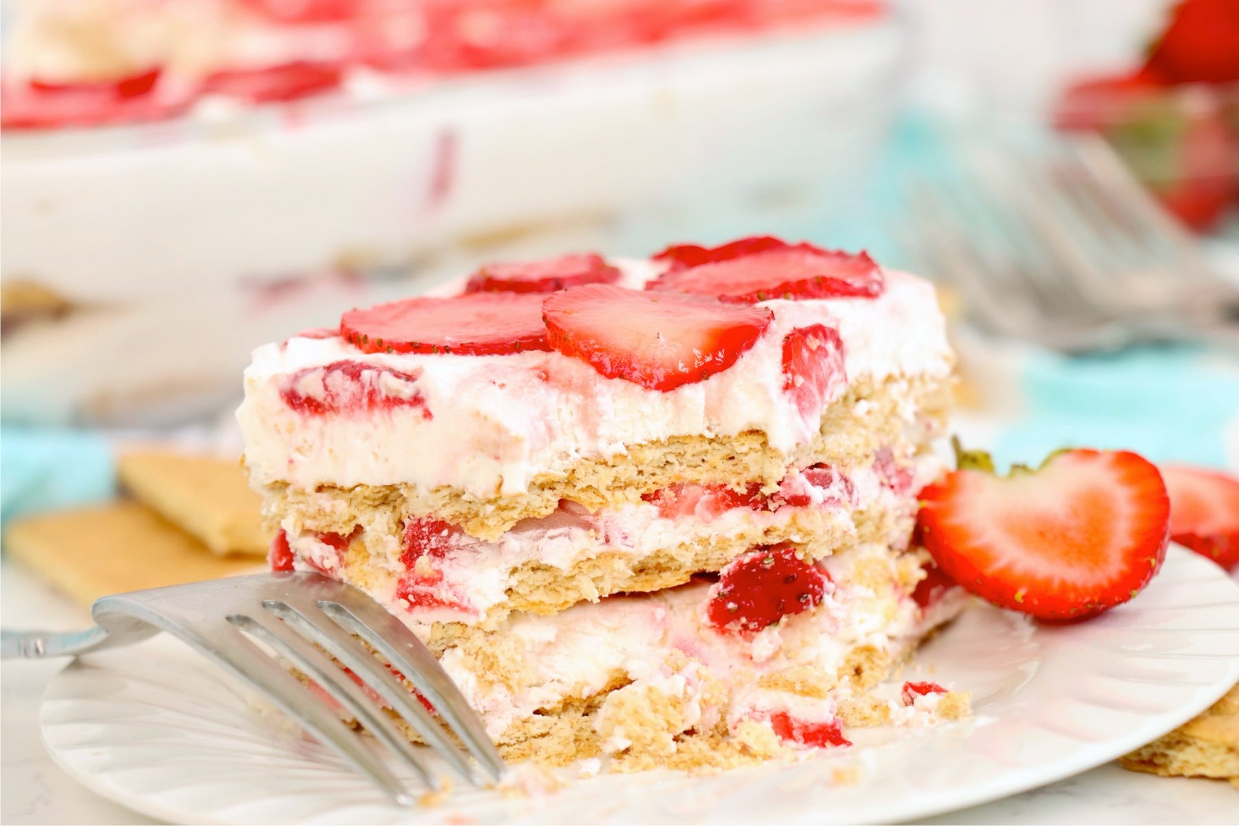 Partially eaten slice of strawberry icebox cake on a plate with a fork and a halved strawberry.