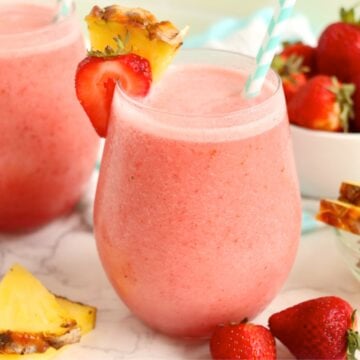 Two pink tropical drinks with pineapple wedges, fresh strawberries, and a bowl of fruit in the background.