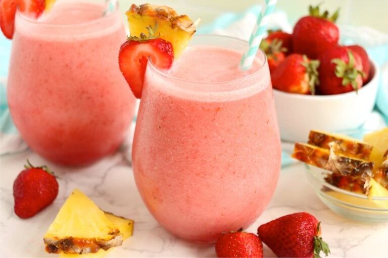 Two pink tropical drinks with pineapple wedges, fresh strawberries, and a bowl of fruit in the background.