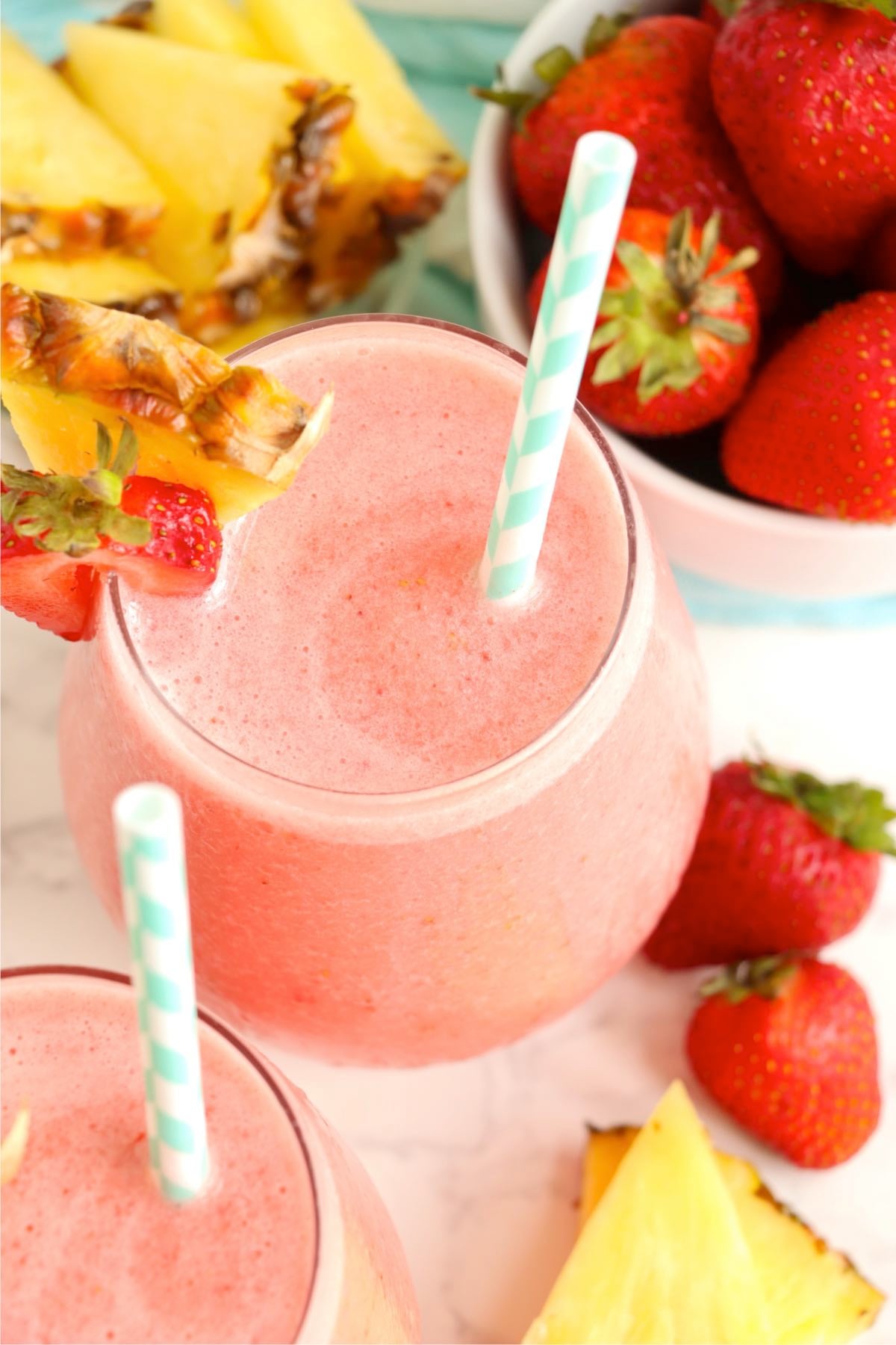 Two glasses of pink tropical drinks with striped straws and fruit garnish.