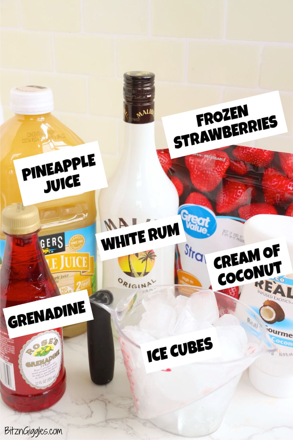 Ingredients on a counter including pineapple juice, white rum, frozen strawberries, cream of coconut, grenadine, and ice cubes.