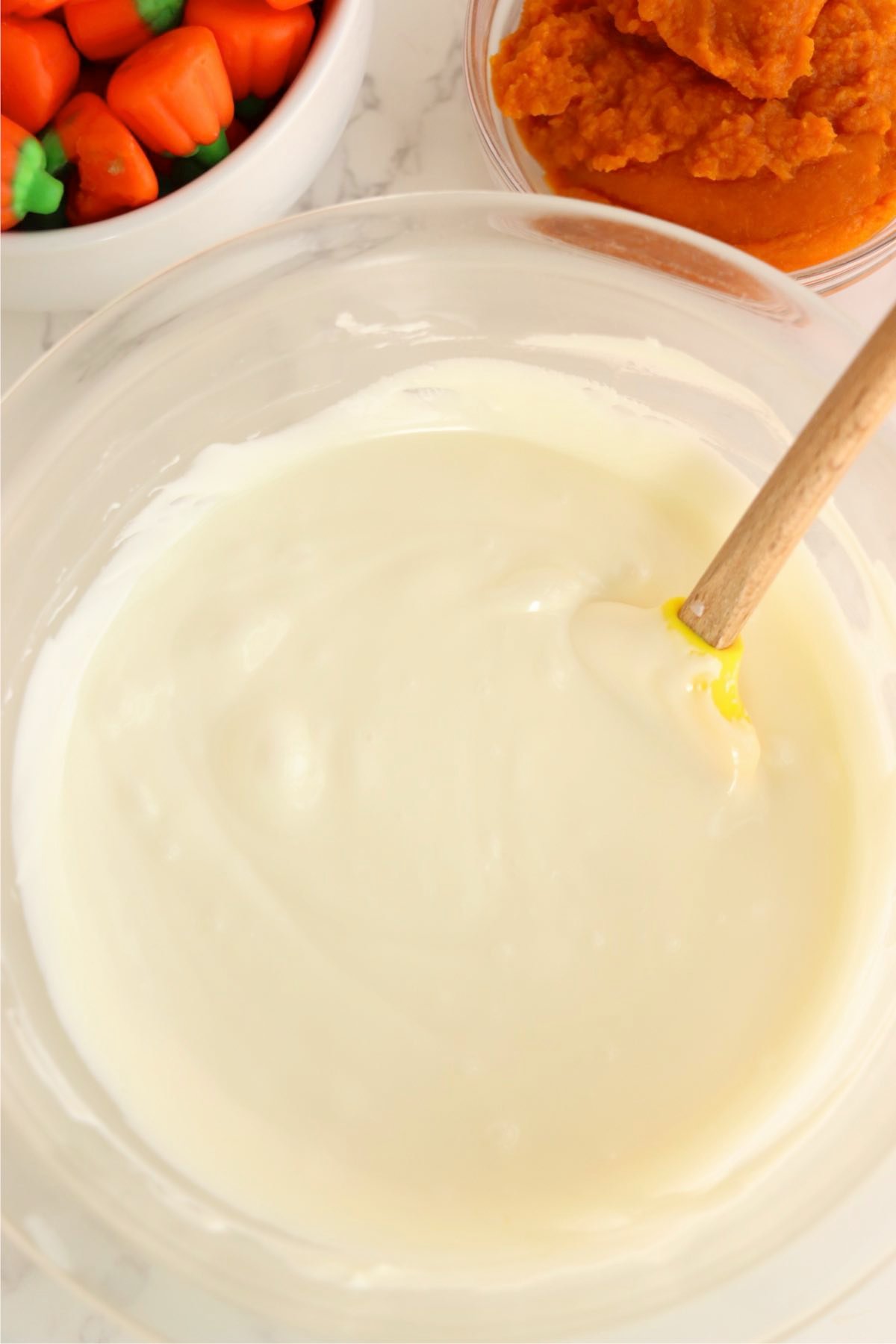 Melted white chocolate in a glass bowl with a spatula beside bowls of pumpkin puree and candy pumpkins.