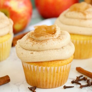 Apple pie stuffed cupcakes topped with cinnamon buttercream and apple filling, surrounded by fresh apples and spices.