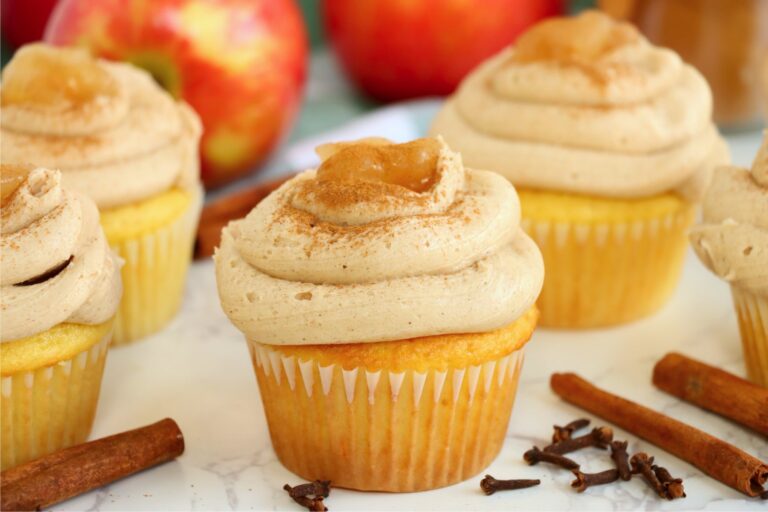 Apple pie stuffed cupcakes topped with cinnamon buttercream and apple filling, surrounded by fresh apples and spices.