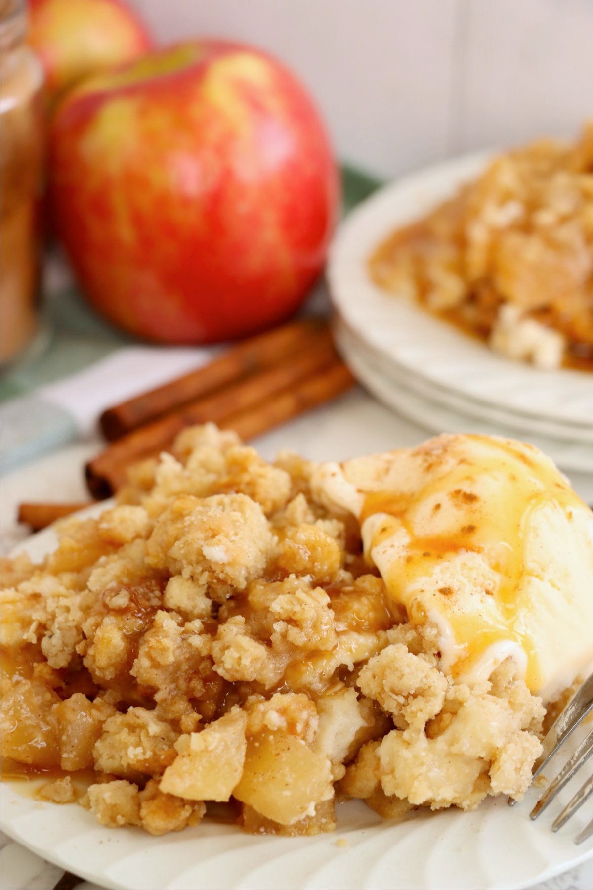 Close-up of a cinnamon apple crisp served with melting ice cream and caramel drizzle, surrounded by fresh apples and cinnamon sticks.