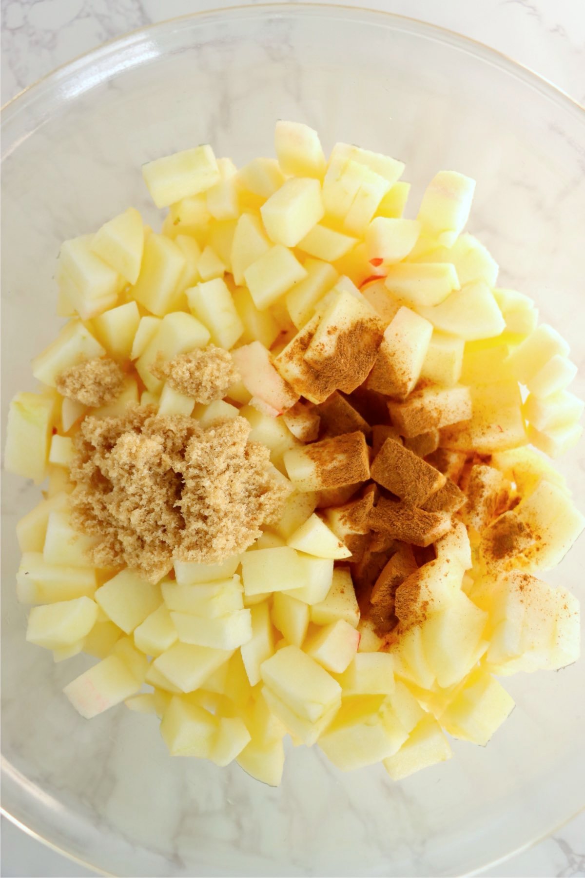 Chopped apples tossed with brown sugar and cinnamon in a glass mixing bowl.