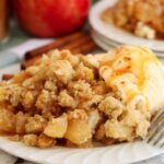 Plate of golden apple crumble with creamy ice cream melting over the top and caramel sauce pooling on the side.