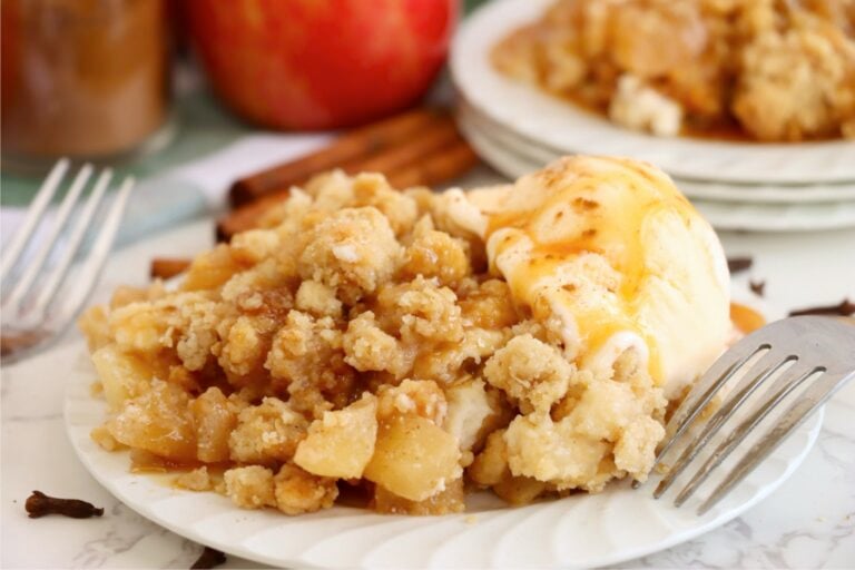 Plate of golden apple crumble with creamy ice cream melting over the top and caramel sauce pooling on the side.