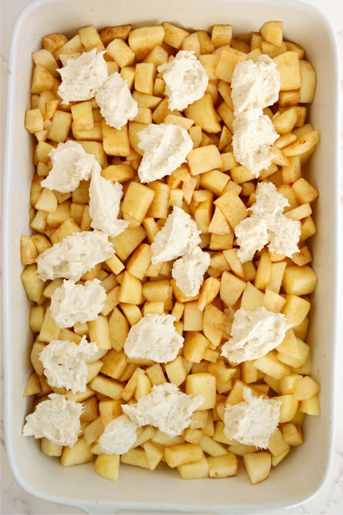 Dollops of creamy cheesecake mixture spooned over spiced apples in a baking dish.