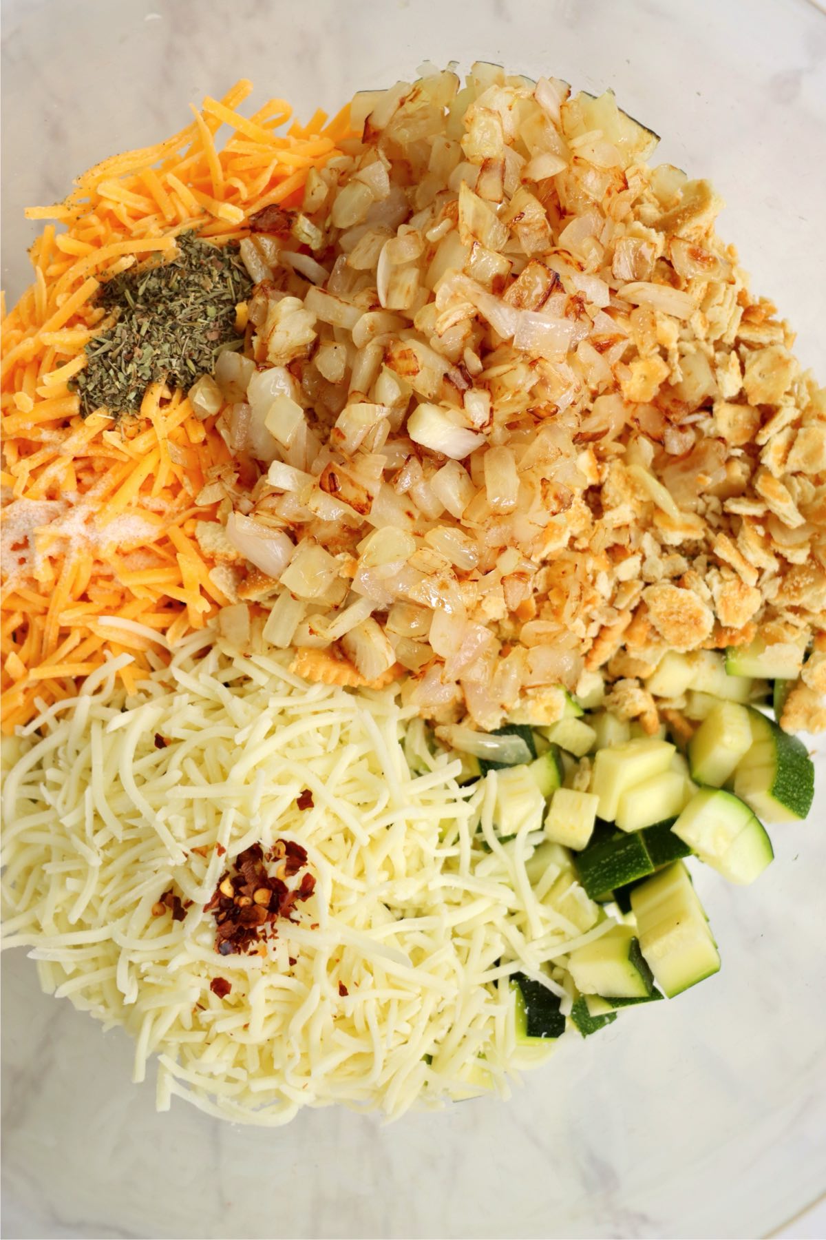 Bowl filled with sautéed onions, shredded cheese, zucchini, herbs, and crushed crackers ready to be mixed.
