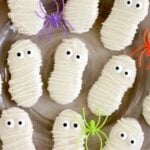 Mummy cookies on a gray plate with pumpkins and plastic spiders in black, purple, orange, and green.