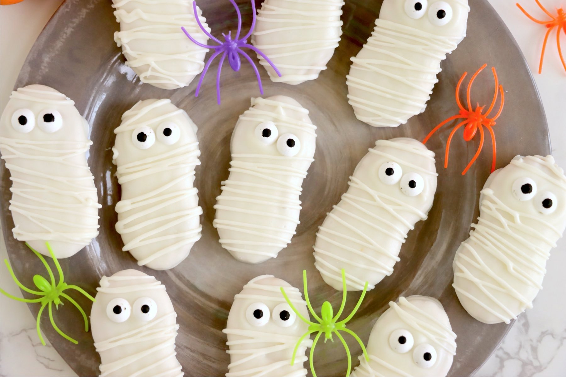Mummy cookies on a gray plate with pumpkins and plastic spiders in black, purple, orange, and green.
