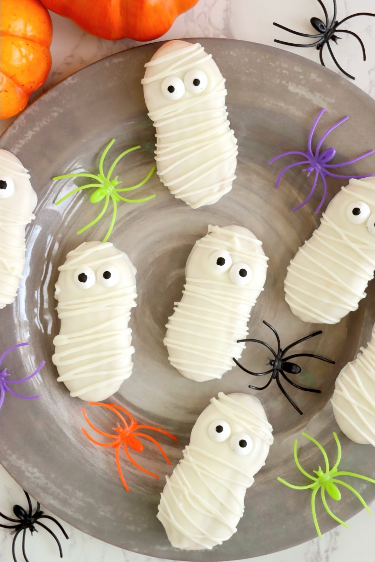 A gray serving plate with several mummy cookies surrounded by colorful plastic spiders.