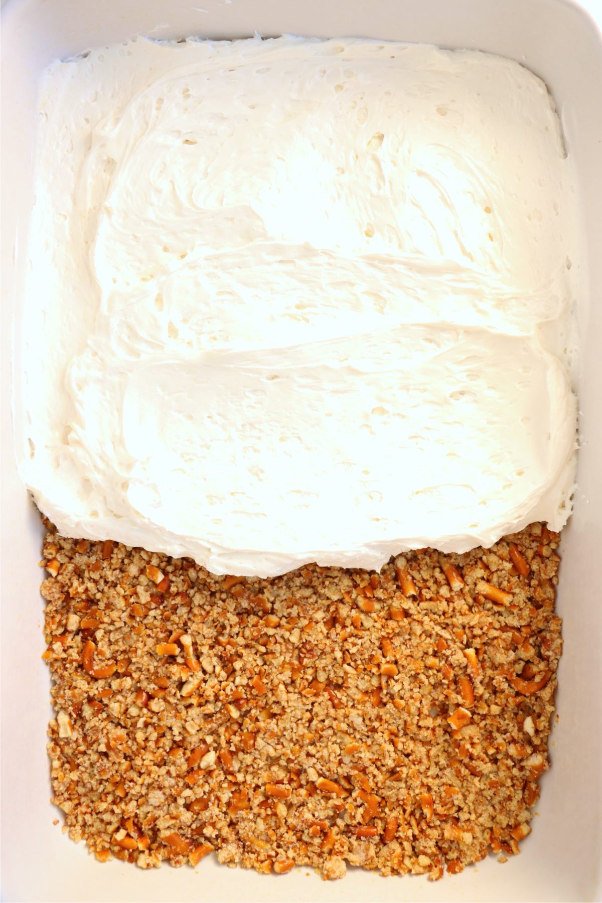 Creamy cheesecake filling being spread evenly over a baked pretzel crust in a dish.