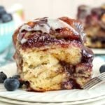 A plated slice of blueberry-filled cinnamon roll with fresh blueberries and a fork.