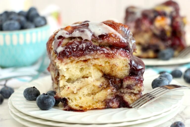A plated slice of blueberry-filled cinnamon roll with fresh blueberries and a fork.