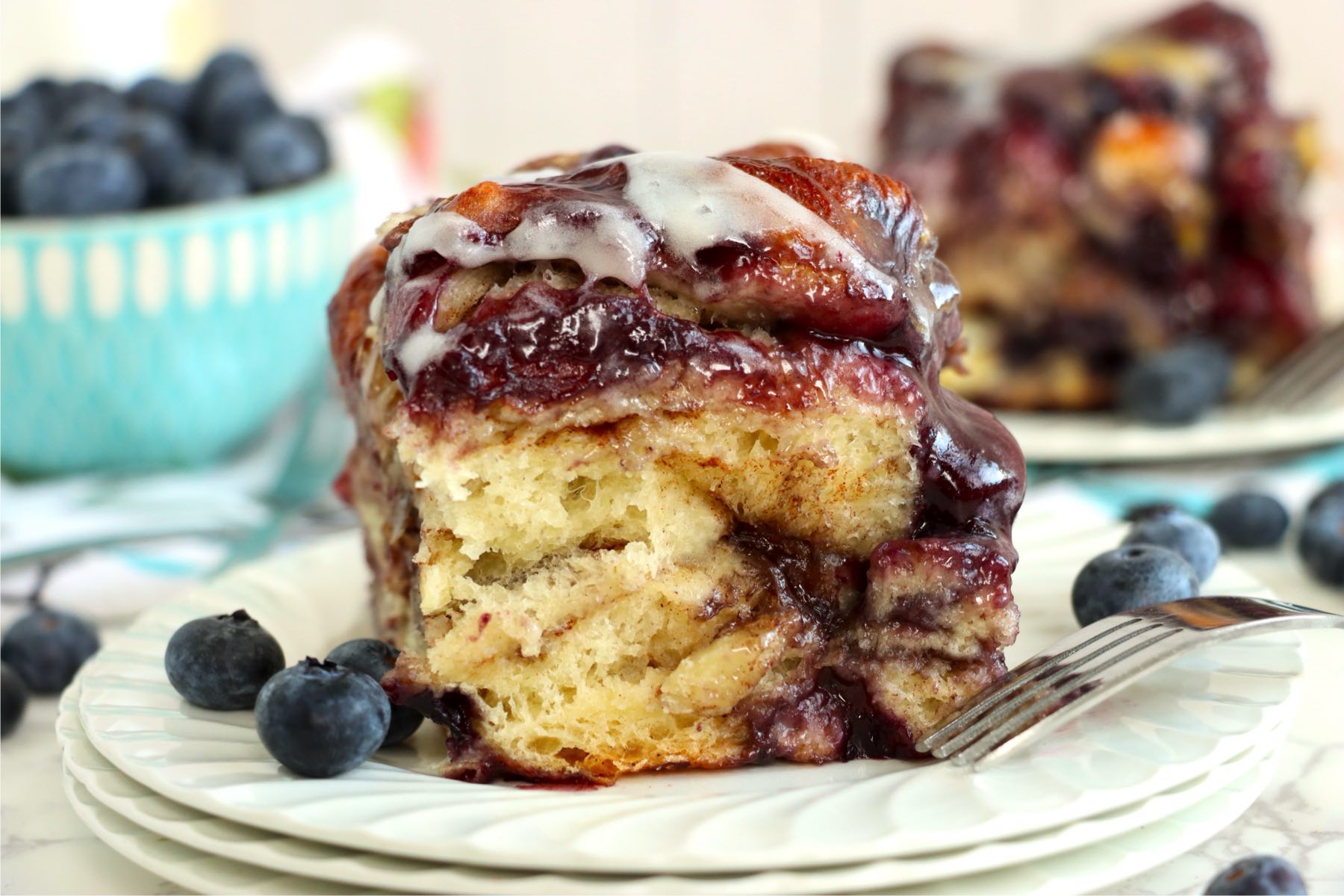 A plated slice of blueberry-filled cinnamon roll with fresh blueberries and a fork.