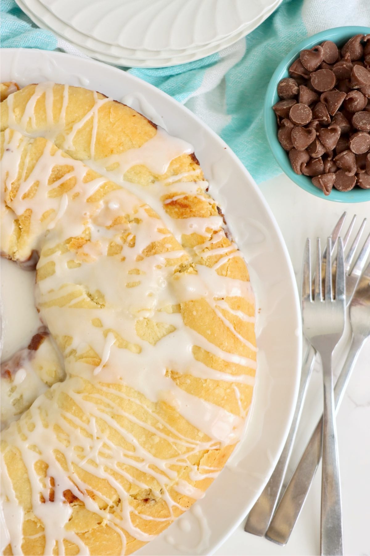 A baked chocolate kringle on a plate with glaze, chocolate chips, and utensils nearby.