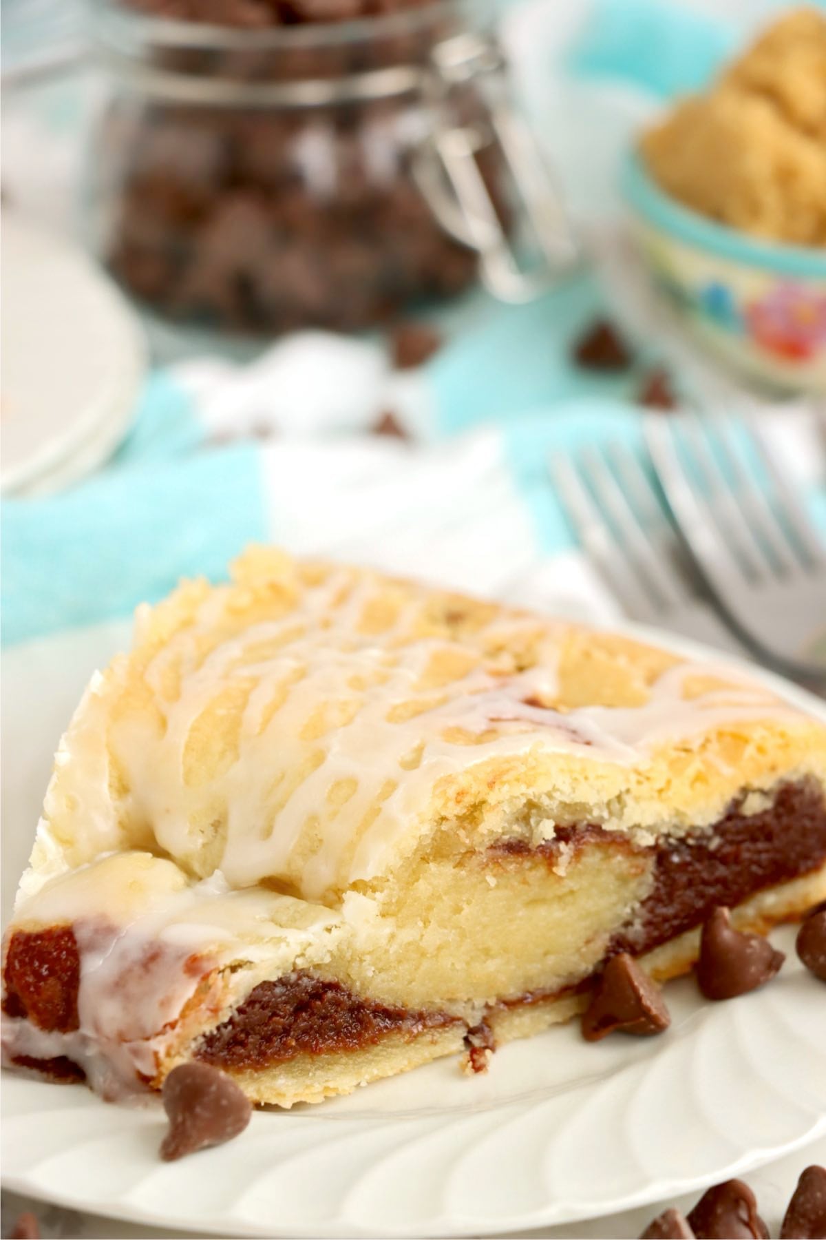 A glazed chocolate kringle slice with forks and ingredients blurred in the background.