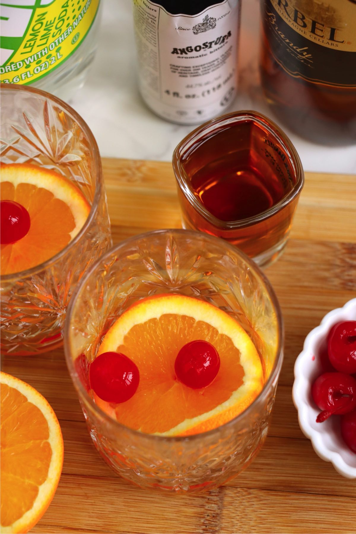 Two cocktail glasses on a wooden board with orange slices, cherries, brandy, and bitters nearby.